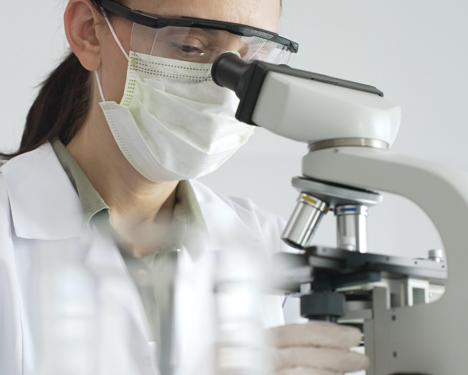 A scientist wearing a face mask and safety goggles looks through a microscope in a lab.