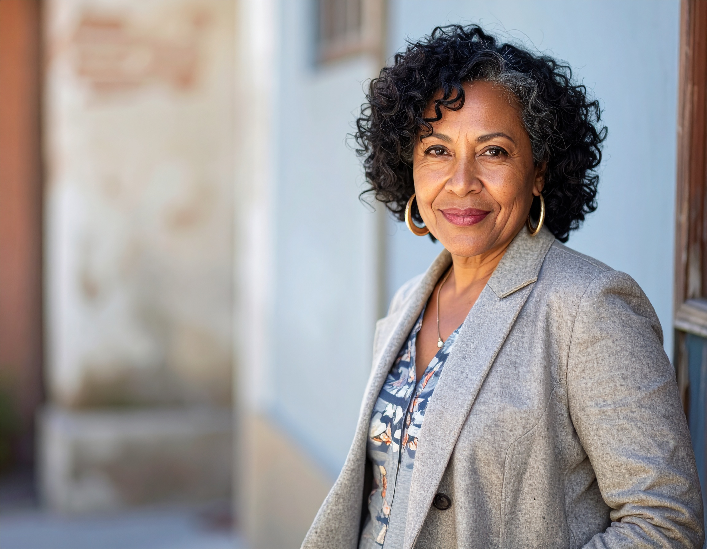 Woman with curly hair in a gray blazer smiling outdoors.