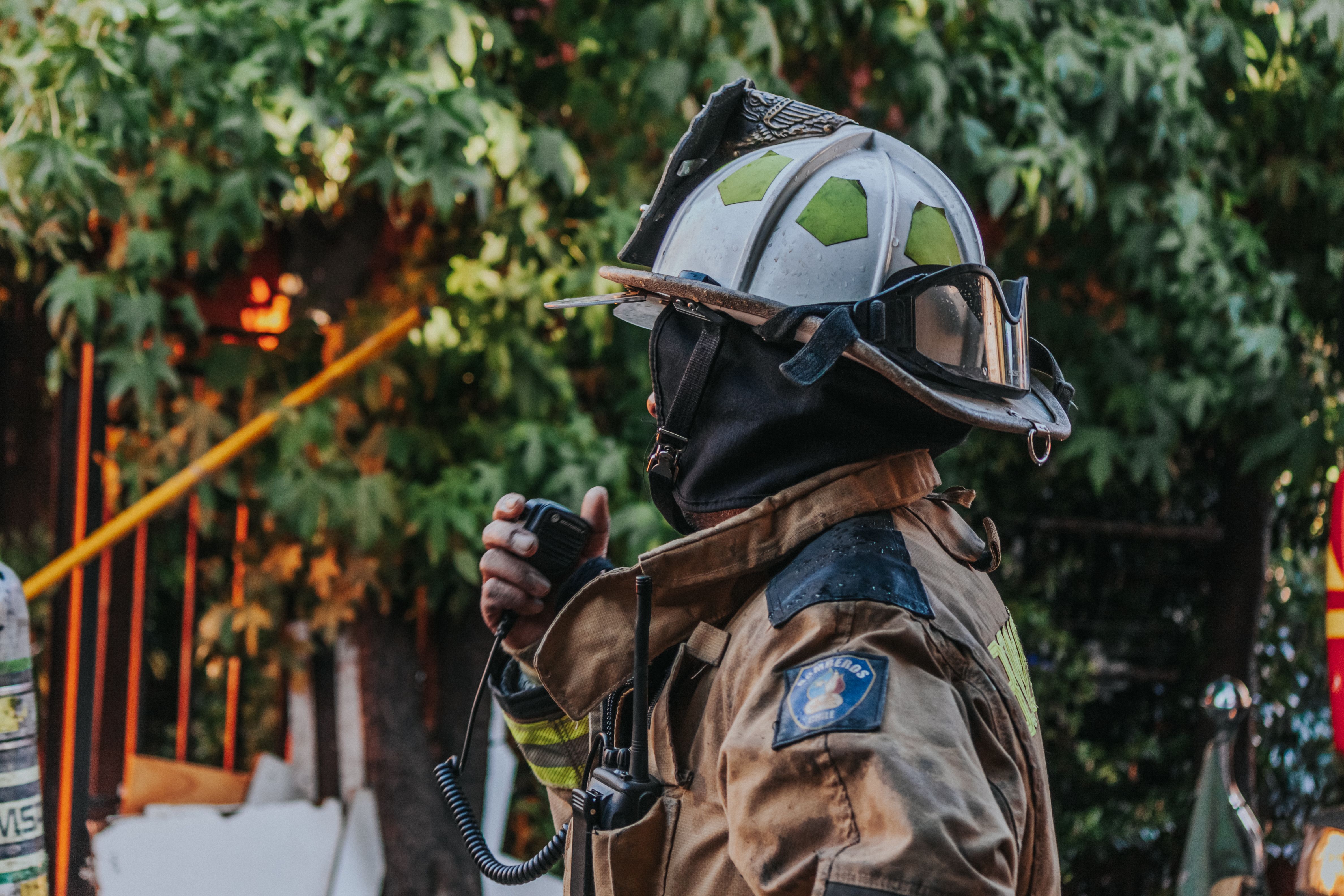 A firefighter in full gear, seen from the side, speaks into a handheld radio with trees behind.
