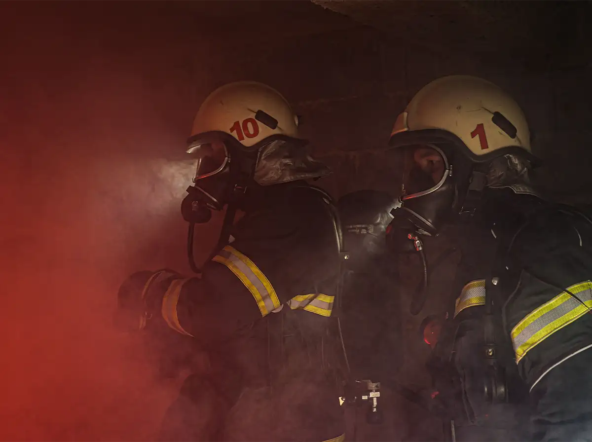 Two firefighters in full gear and masks stand in a dark room filled with red and white smoke.
