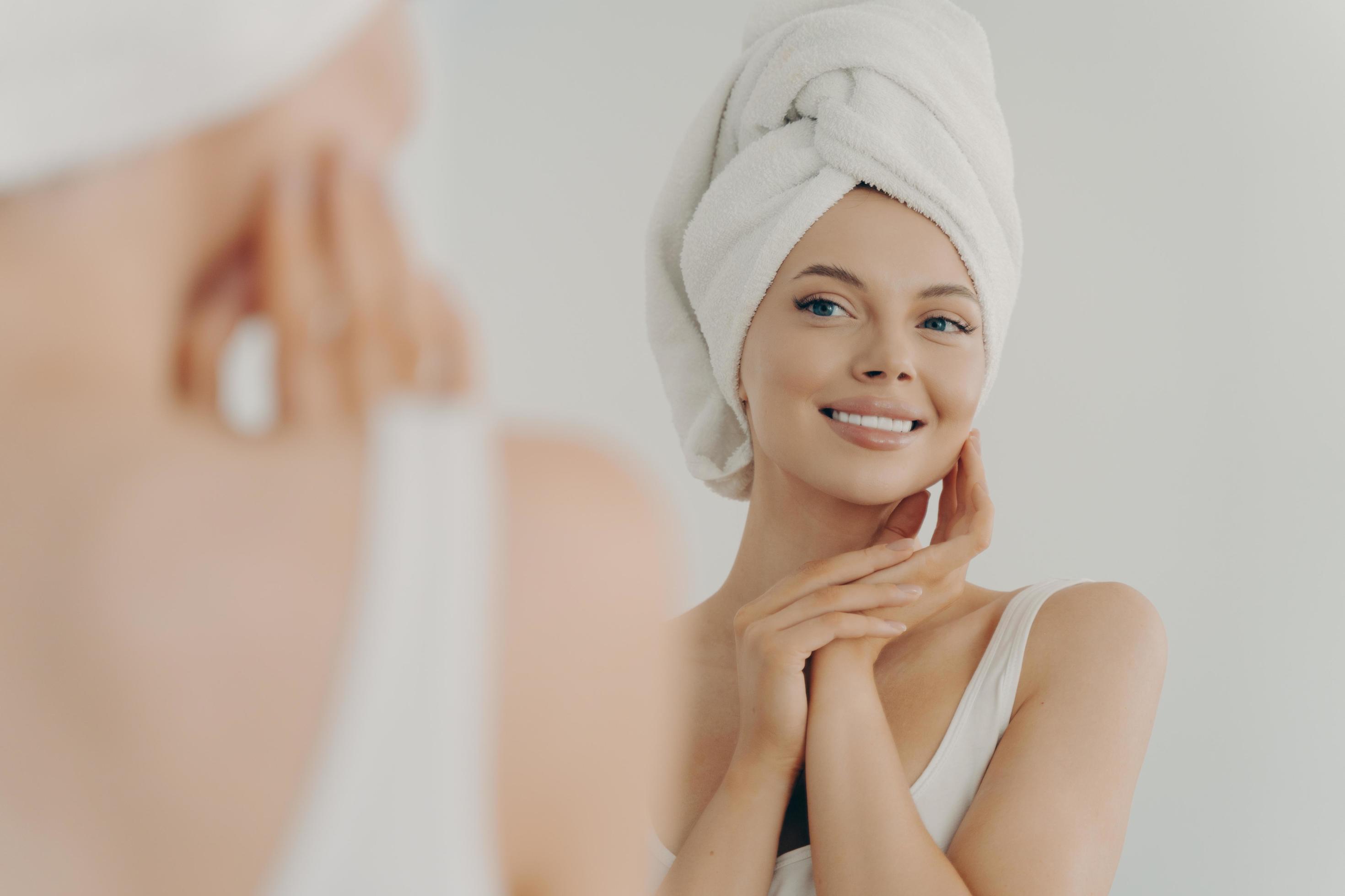 Woman with towel on head, smiling in mirror.