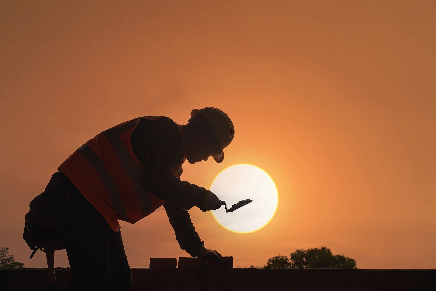 A silhouette of a construction worker wearing a hard hat and laying bricks against the setting sun.