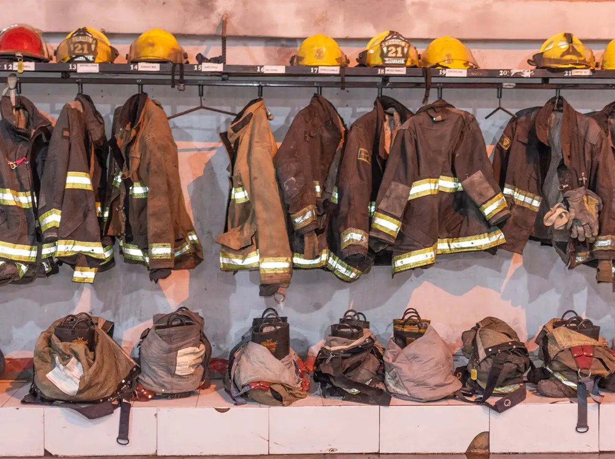 Worn firefighter jackets hang on a rack below a shelf of helmets in a fire station locker room.