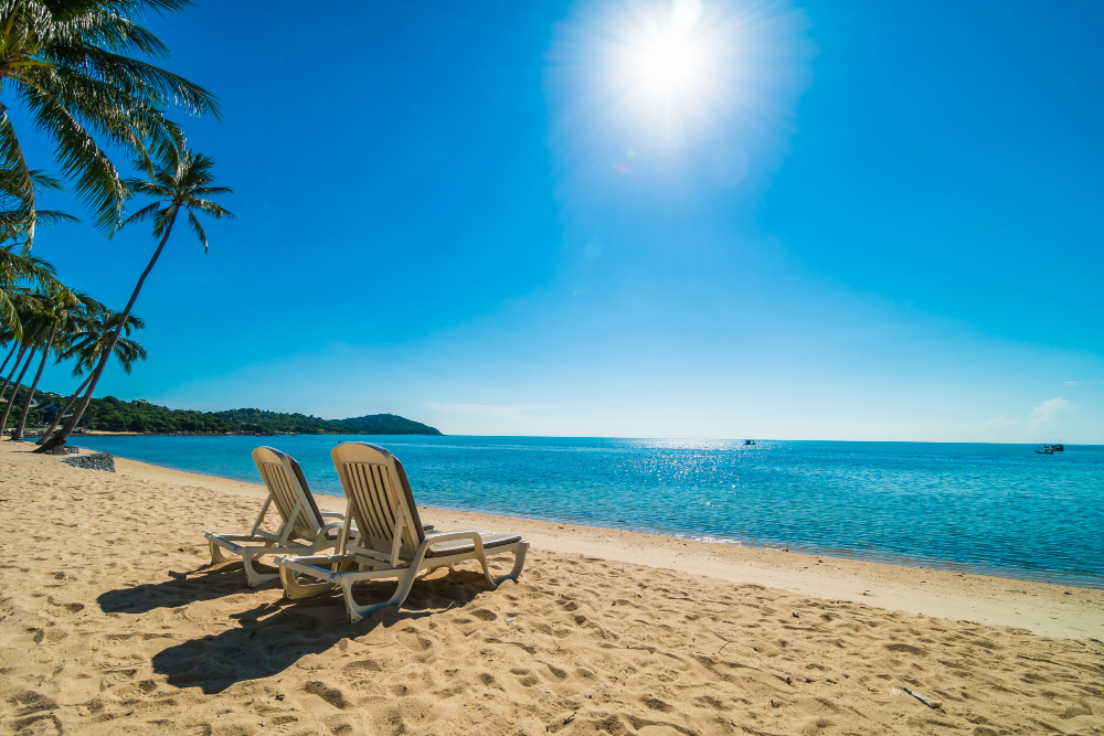 Two empty lounge chairs on a sandy beach facing a calm blue ocean under a bright sun.