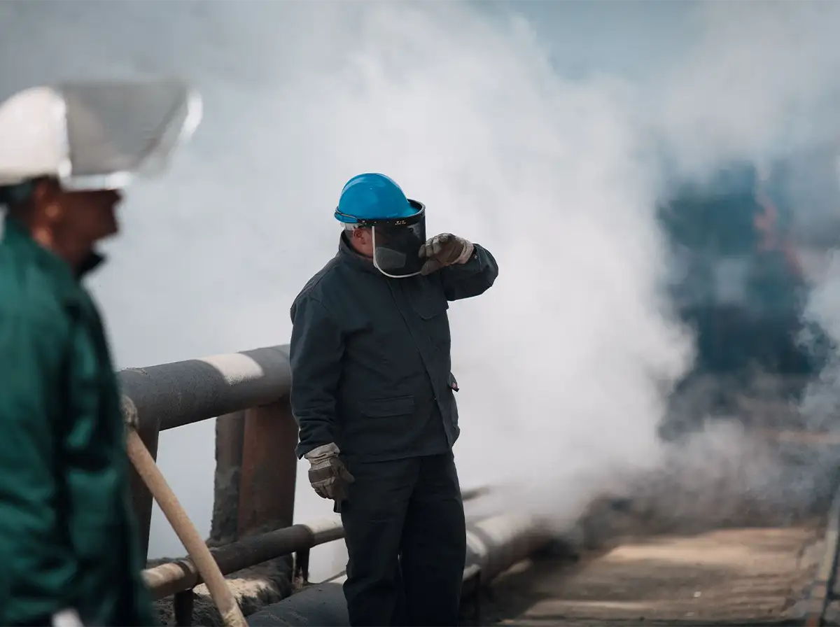 An industrial worker in a blue hard hat and face shield adjusts his visor amid clouds of steam.