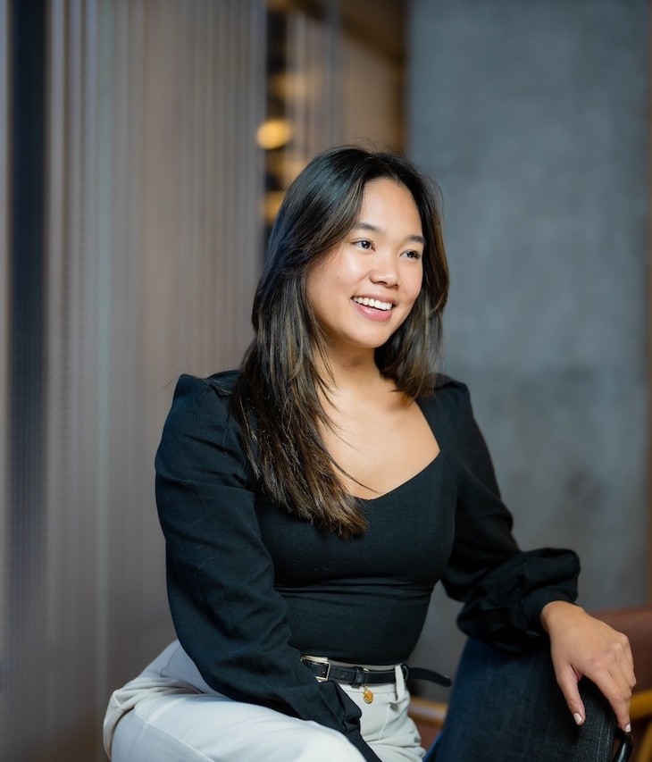 Person seated and smiling in a black top, with a blurred background.