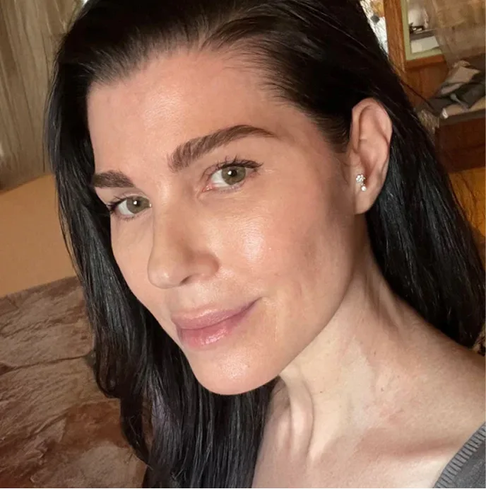 A close-up selfie of a woman with long dark hair and light green eyes looking at the camera.