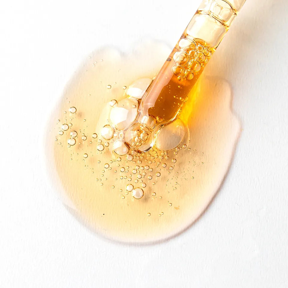A close-up of a glass dropper dispensing a bubbly, golden liquid onto a white surface.