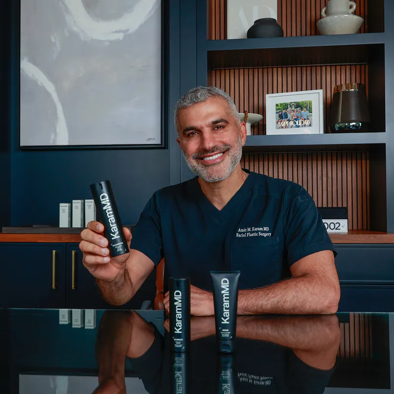 Man in a blue scrub holds KaramMD skincare product, smiling at a desk with shelves behind.
