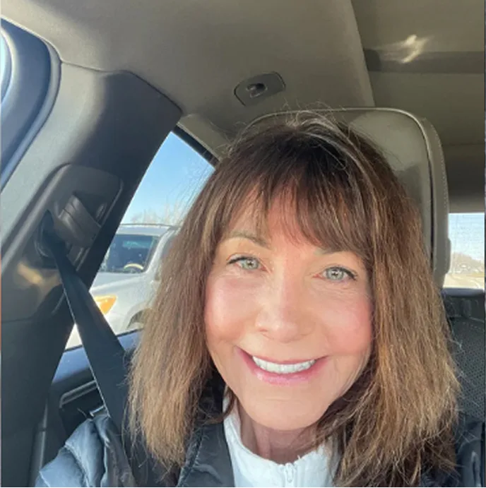 A smiling woman with brown, shoulder-length hair and bangs takes a selfie from the driver's seat of a car.