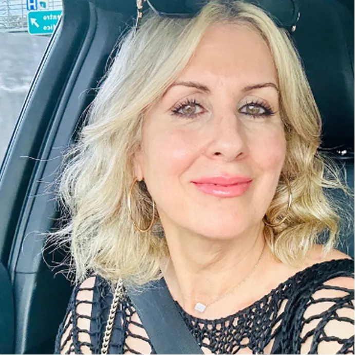 A blonde woman with hoop earrings and a black crochet top smiles while sitting in a car.