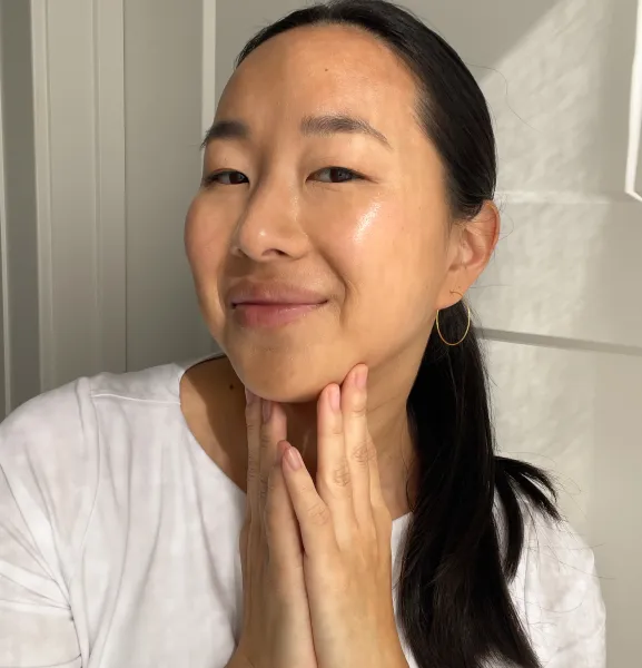 A smiling woman in a white t-shirt gently holds her hands to her chin and neck.