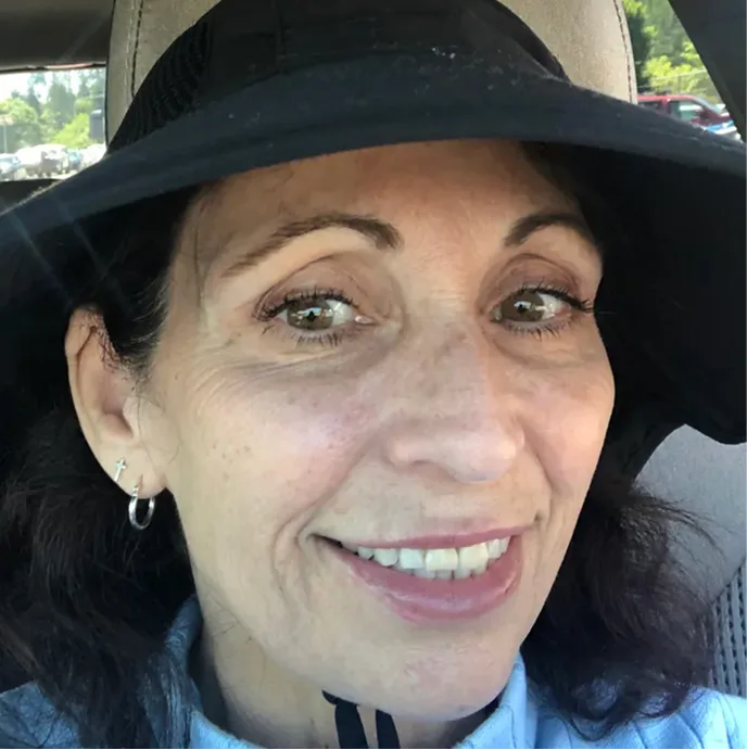 A smiling woman with dark hair wearing a wide-brimmed black hat, taking a selfie in a car.