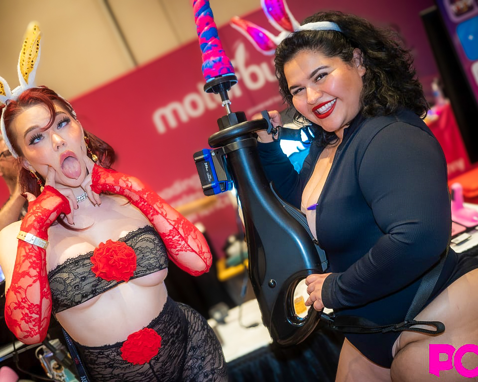 Two women in costumes pose at an event, one holding a large machine with a sex toy.