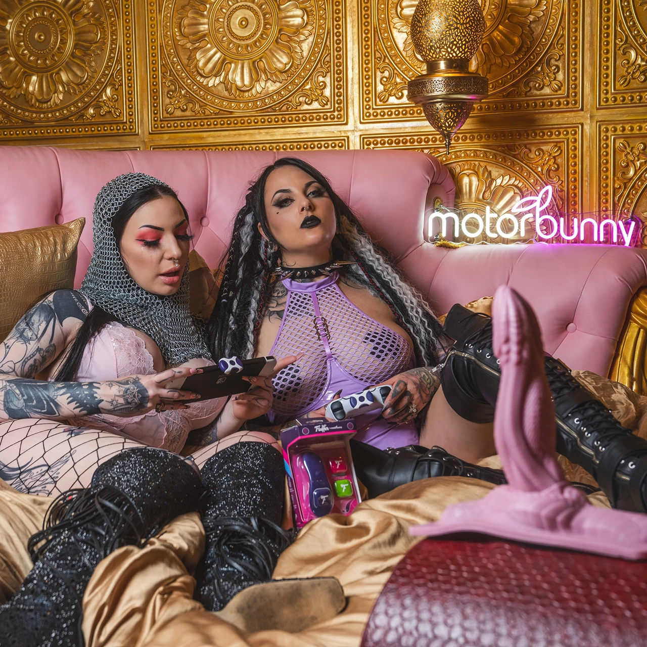
Two tattooed alternative women sitting on a pink couch with gold bedding, in alternative lingerie - with a Motorbunny in the forefront and Fluffer enabled toys between them