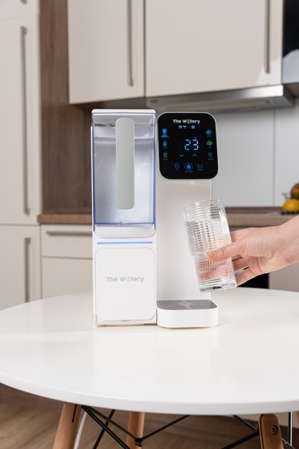 Person filling a glass with water from a countertop water dispenser.