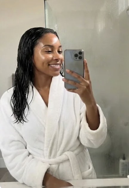 A smiling woman with wet hair, wearing a white bathrobe, takes a selfie in a mirror.