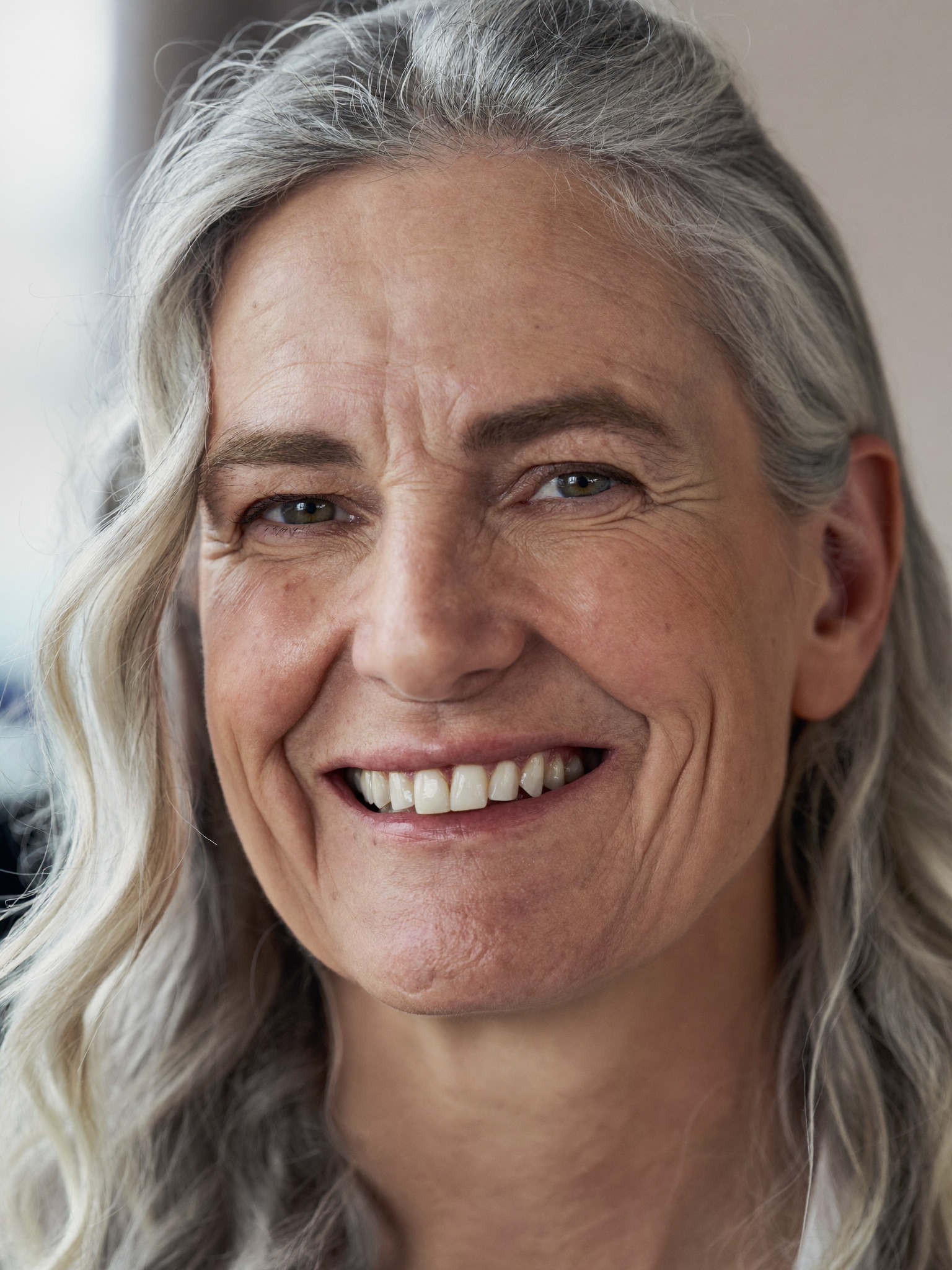 A close-up portrait of a smiling older woman with long, gray hair looking directly at the camera.