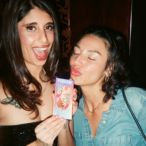 Two women posing together; one holds up a small box while the other makes a kissing face at it.