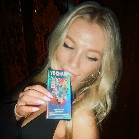 A blonde woman with winged eyeliner holds a colorful chocolate bar that says 'Yeehaw' to her lips.