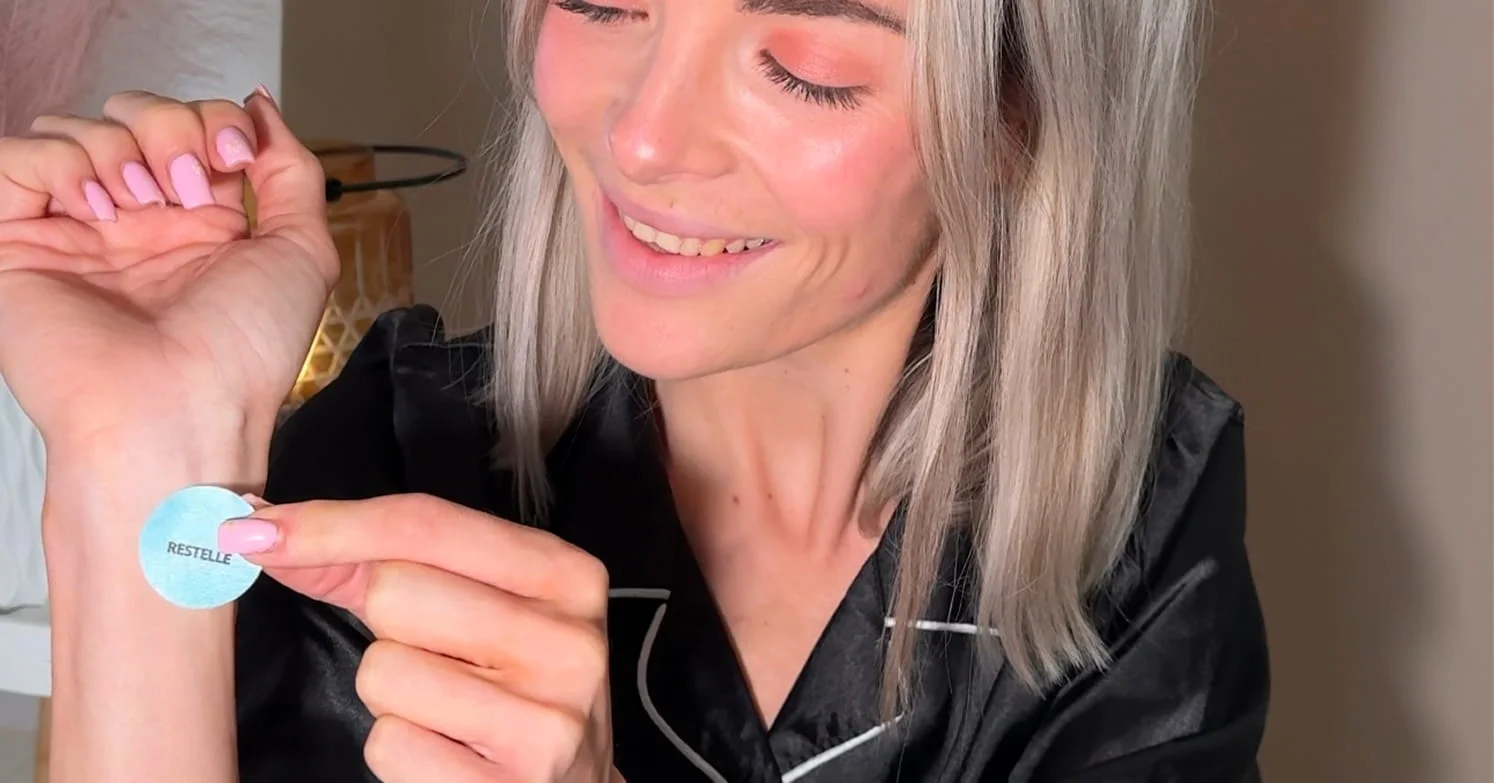 A smiling blonde woman applies a small, round, blue patch labeled 'RESTELLE' to her inner wrist.