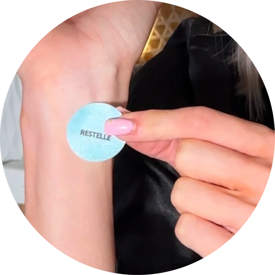 A close-up of a hand with pink nails applying a round, blue 'Restelle' patch to a wrist.