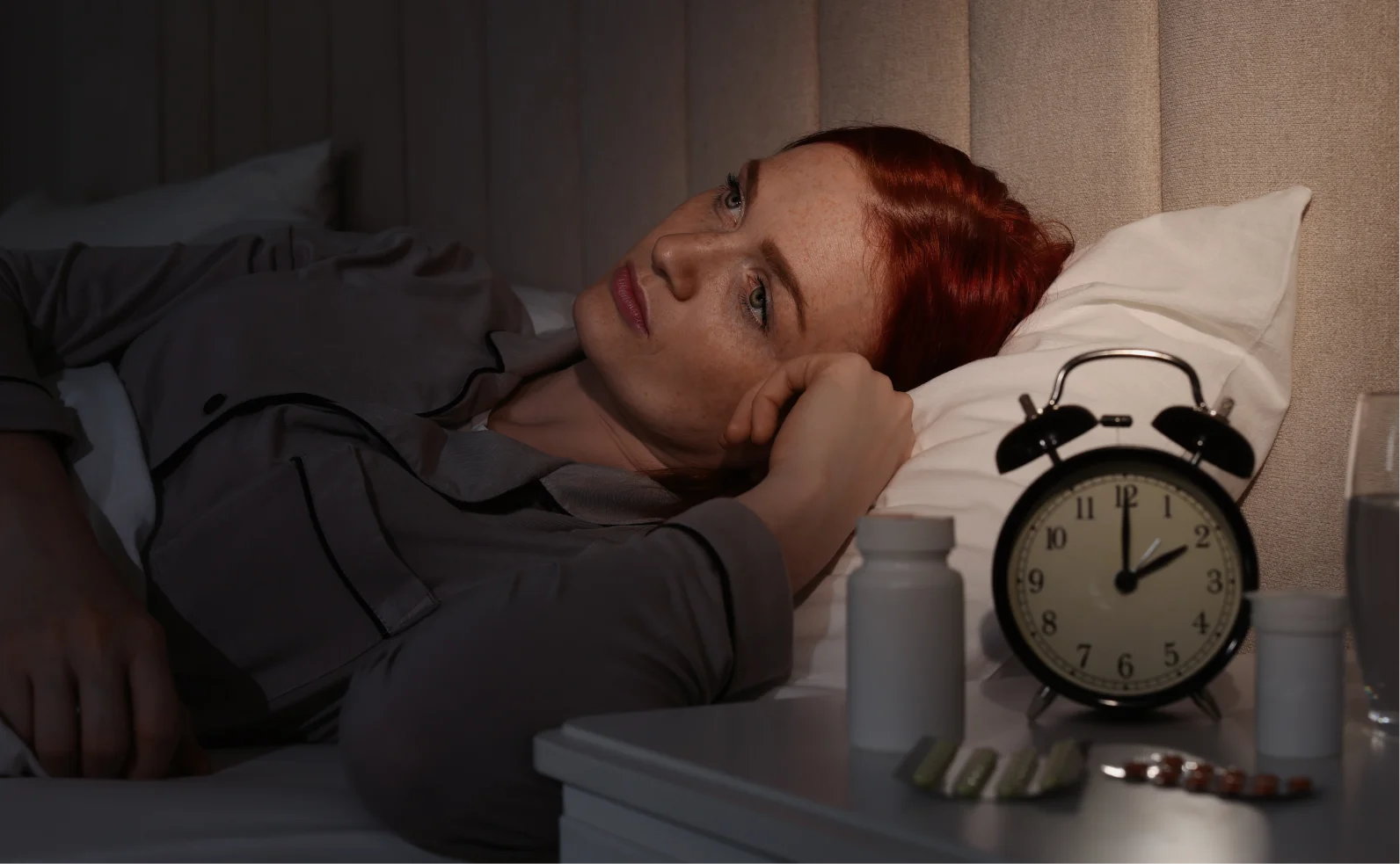 A woman with red hair lies awake in bed with an alarm clock and medication on her nightstand.