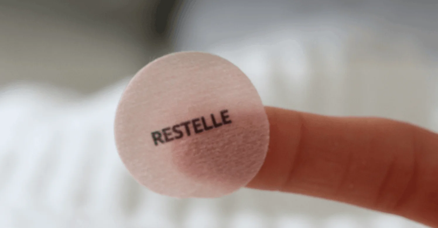 A close-up of a fingertip holding a small, round, translucent patch with the word RESTELLE on it.
