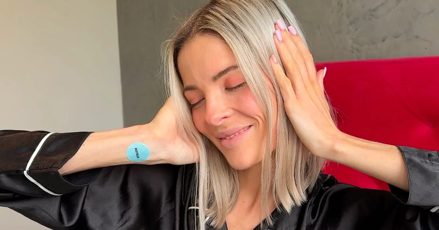 A smiling blonde woman in black pajamas with eyes closed and a blue patch on her wrist.