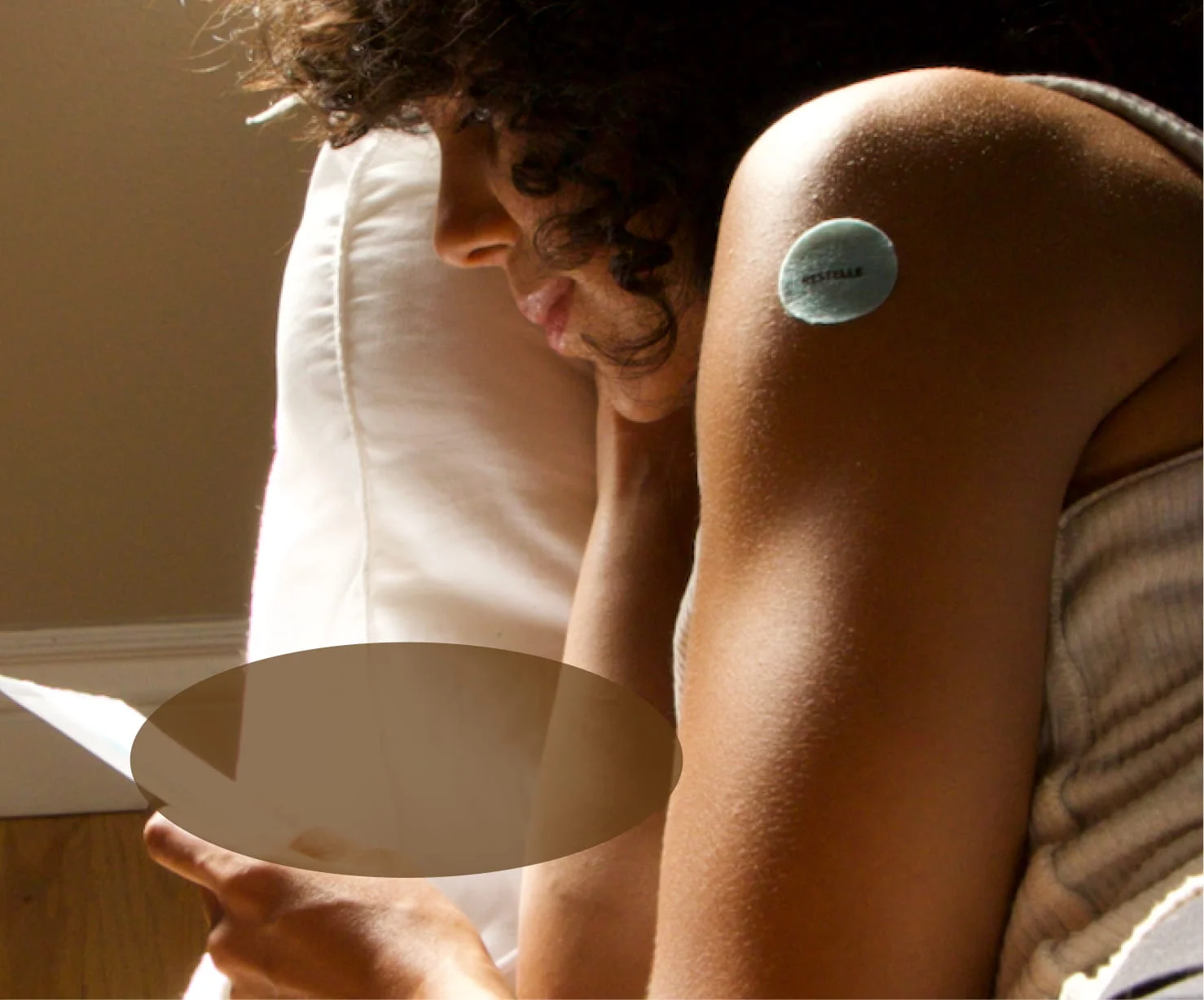 A person with a round patch on their arm rests their head on a pillow in the sunlight.