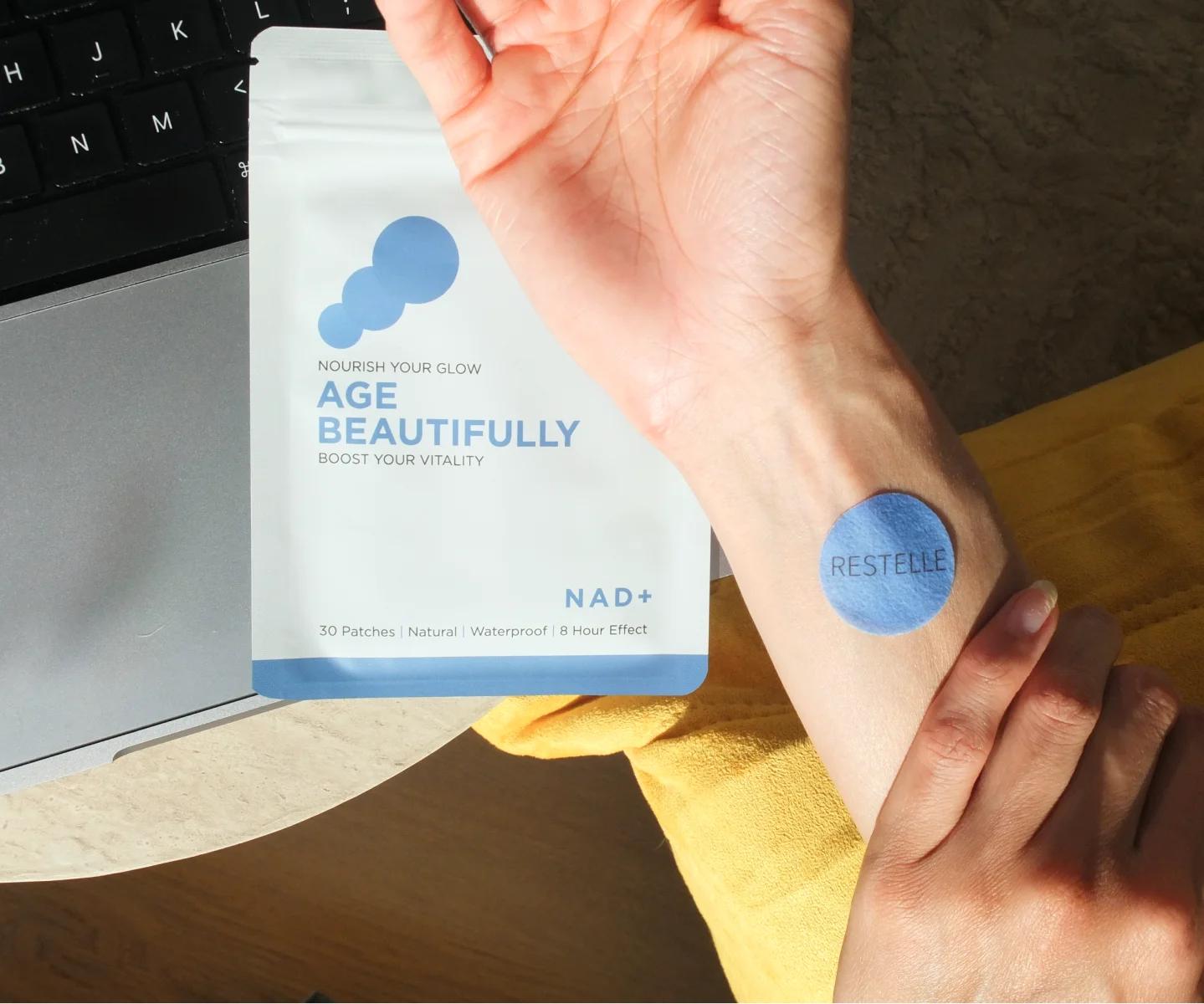 A person's wrist with a blue Restelle patch, next to its 'Age Beautifully' packaging and a laptop.