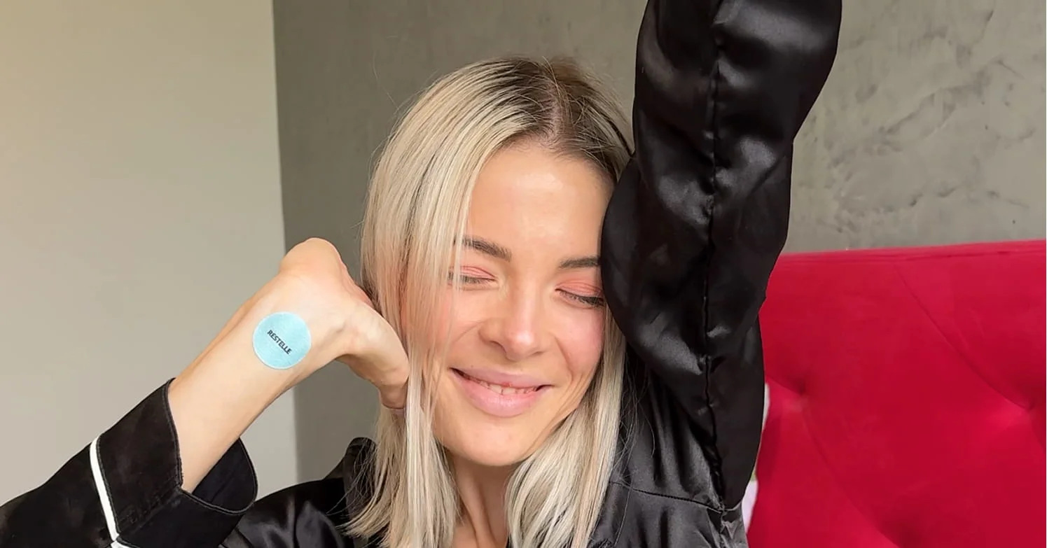 A smiling blonde woman in a black satin top has a blue circular patch on the back of her hand.