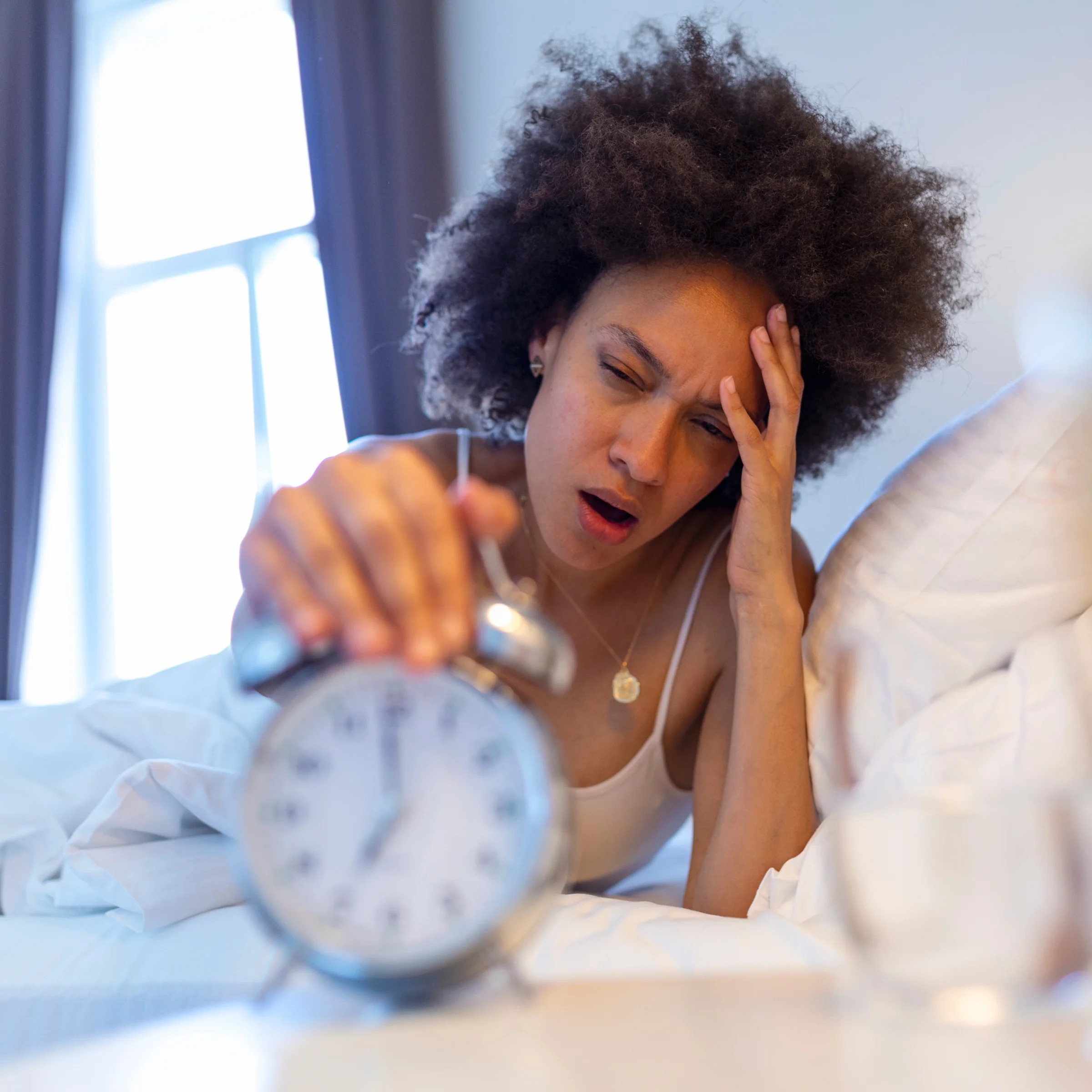A tired-looking woman in bed reaches out to turn off her silver alarm clock.