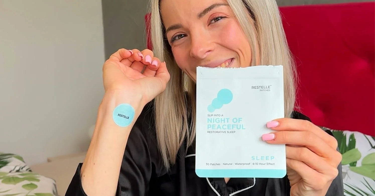 A smiling woman holds a packet of Restelle sleep patches while wearing one on her wrist.