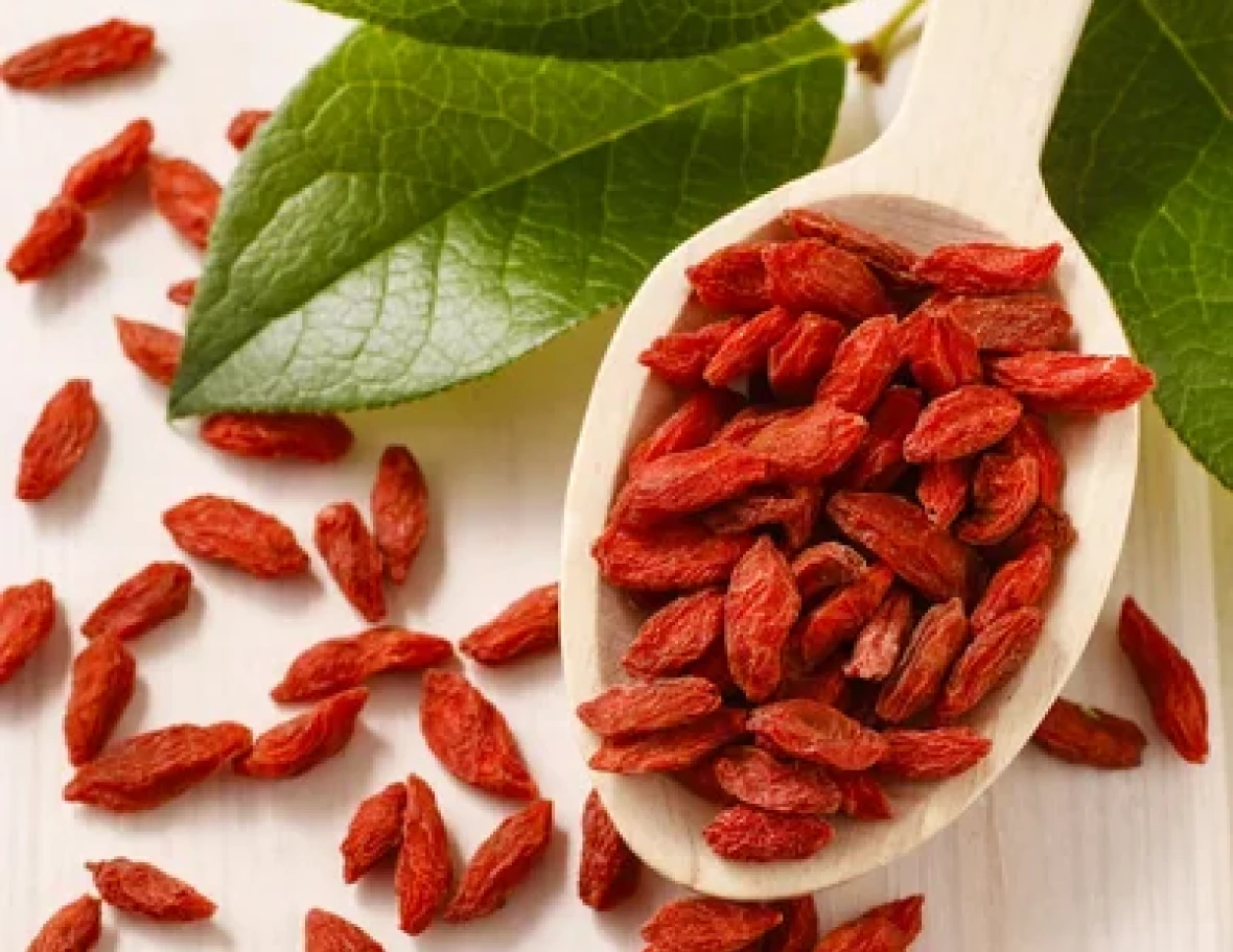 Red dried goji berries on a wooden spoon with green leaves.