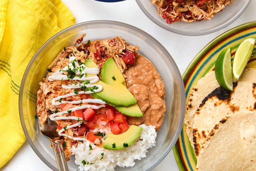 An overhead shot of a chicken burrito bowl with rice, beans, avocado, tomatoes, and a side of tortillas.