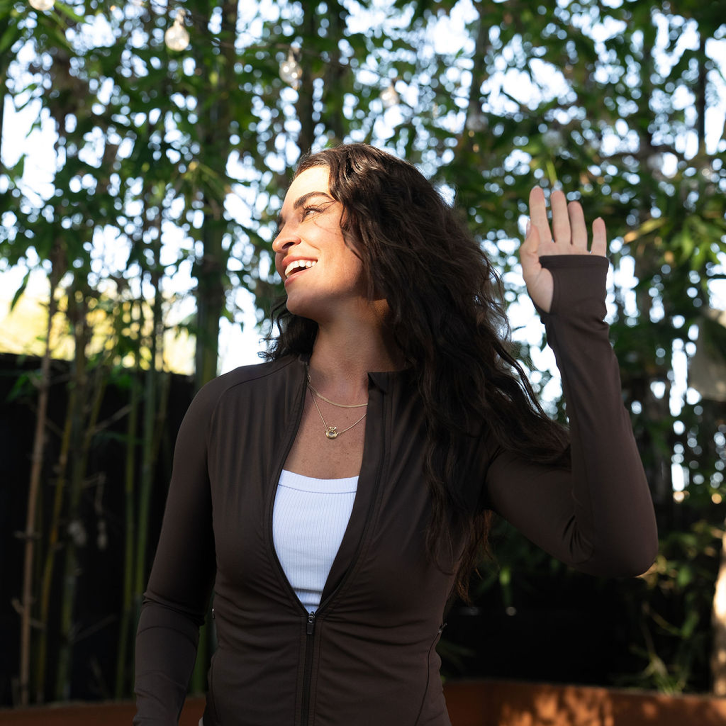 A woman in a brown athletic jacket smiles and waves while standing in front of bamboo trees.