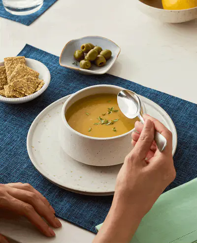 A person eating a bowl of soup, with crackers and olives on the side on a blue placemat.
