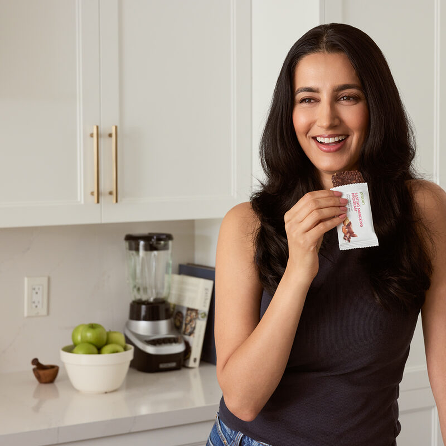 A smiling woman in a kitchen holds a partially unwrapped nutrition bar, about to take a bite.
