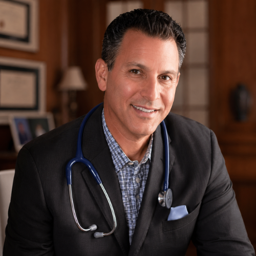 A smiling male doctor in a blazer with a stethoscope around his neck, sitting in an office.