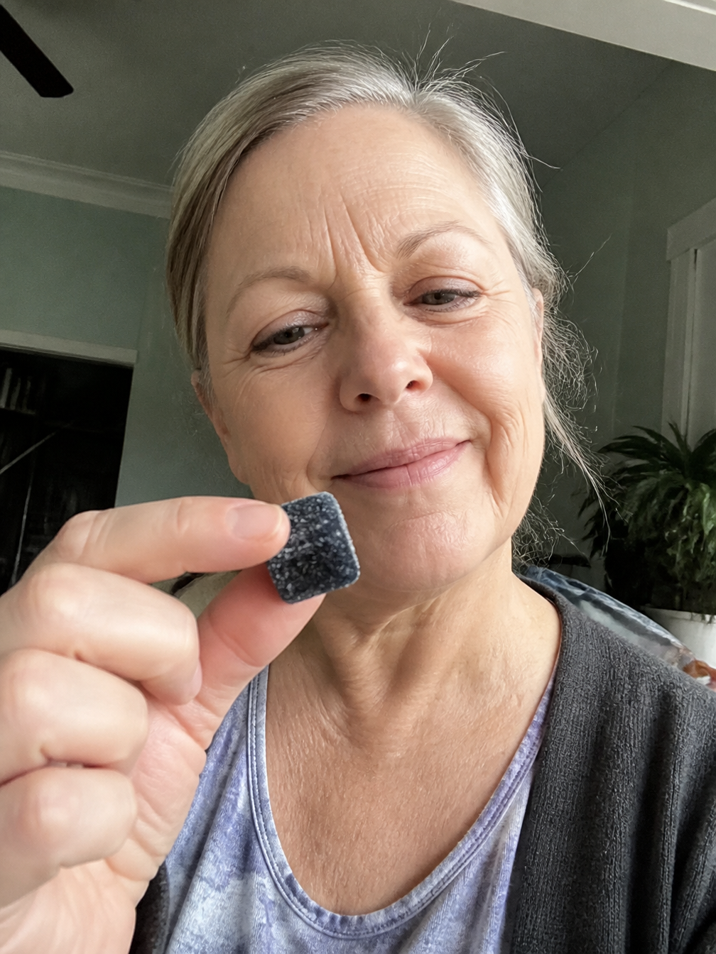 A woman with graying hair smiles while holding up a small, dark, square gummy.