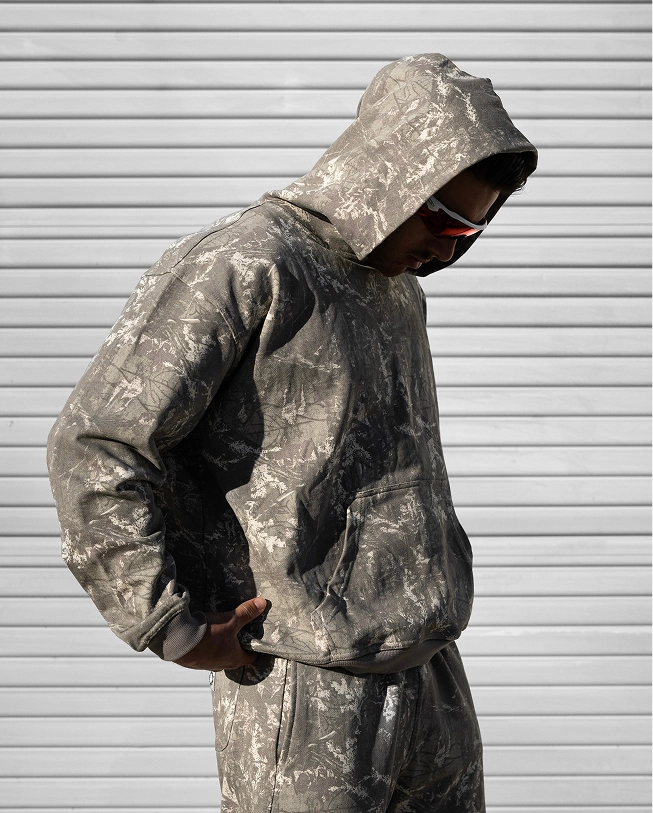 Person in a camo hoodie and pants, wearing sunglasses, against a striped background.