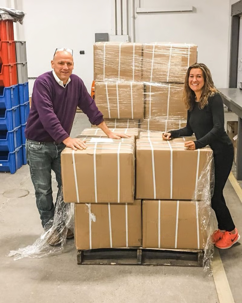 A man and a woman stand on either side of a pallet stacked with cardboard boxes in a warehouse.