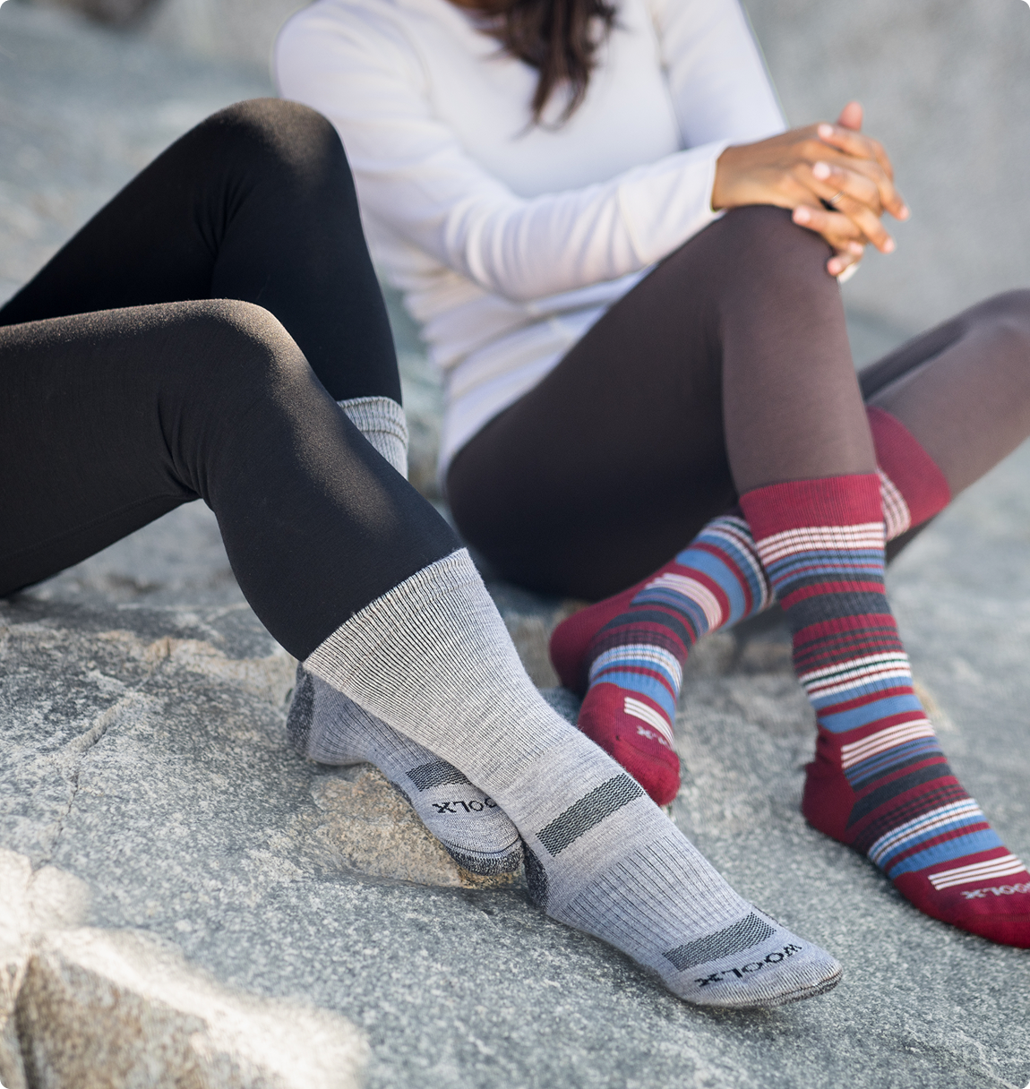 Two people wearing leggings and different pairs of crew socks sit on a large grey rock.