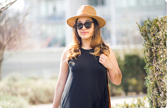 Woman in sunglasses and hat standing outdoors, wearing a black dress.