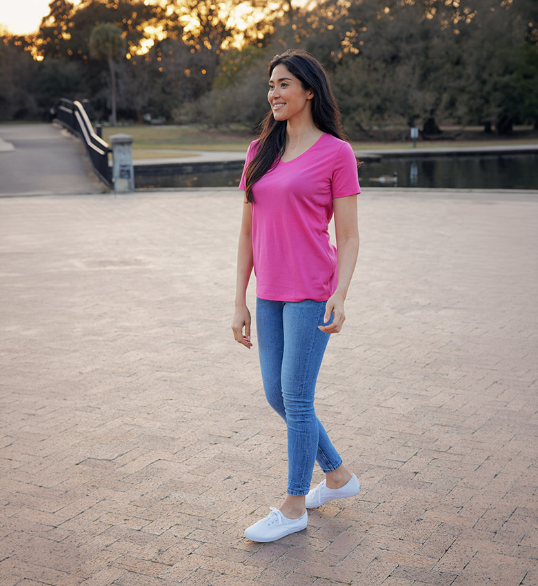 A woman in a pink t-shirt, blue jeans, and white sneakers walking on a paved path in a park.