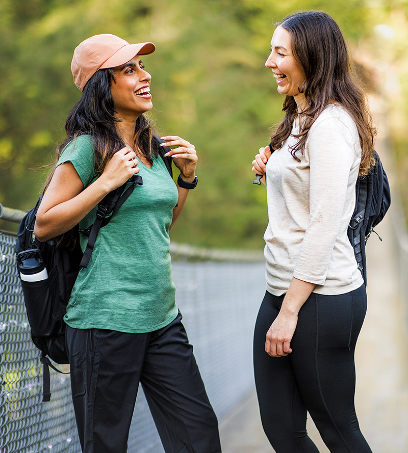 Two women wearing backpacks and athletic clothes stand on a bridge, smiling and talking to each other.