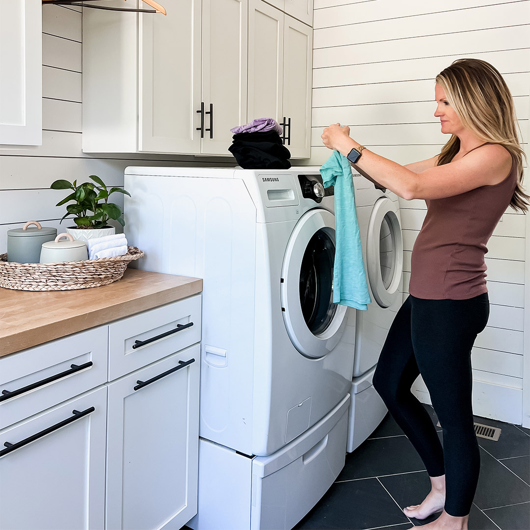 A woman stands in a modern laundry room, loading a shirt into a front-load washing machine.
