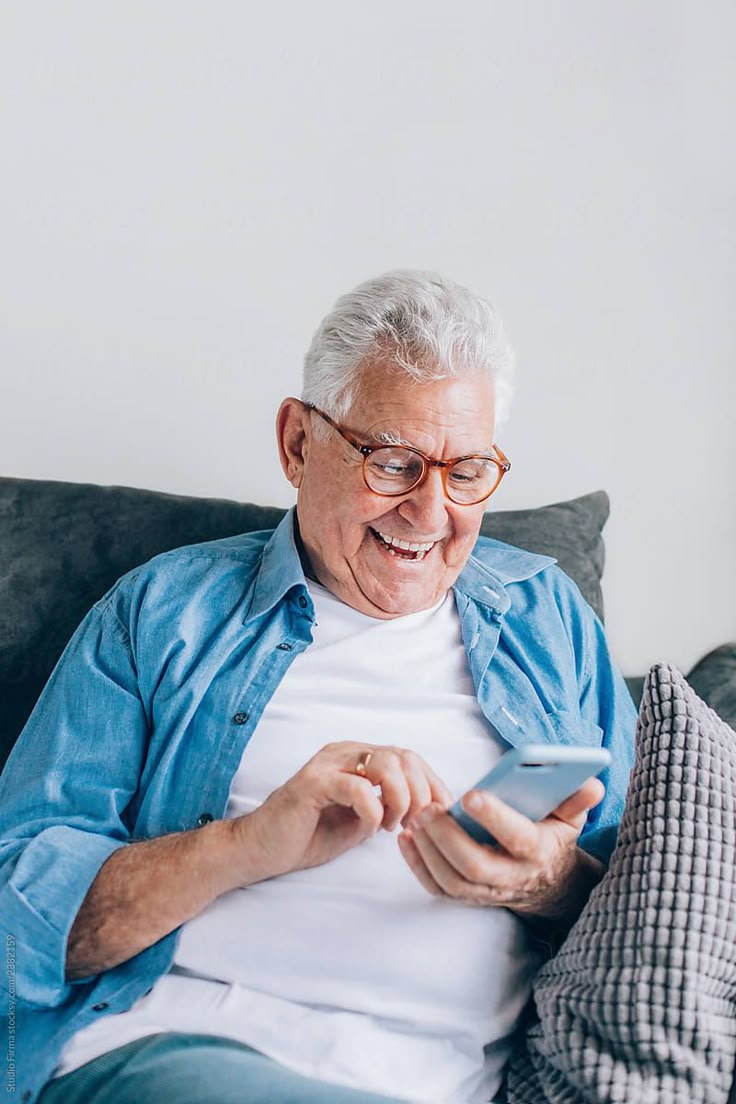 Elderly man smiling while using a smartphone on a couch.
