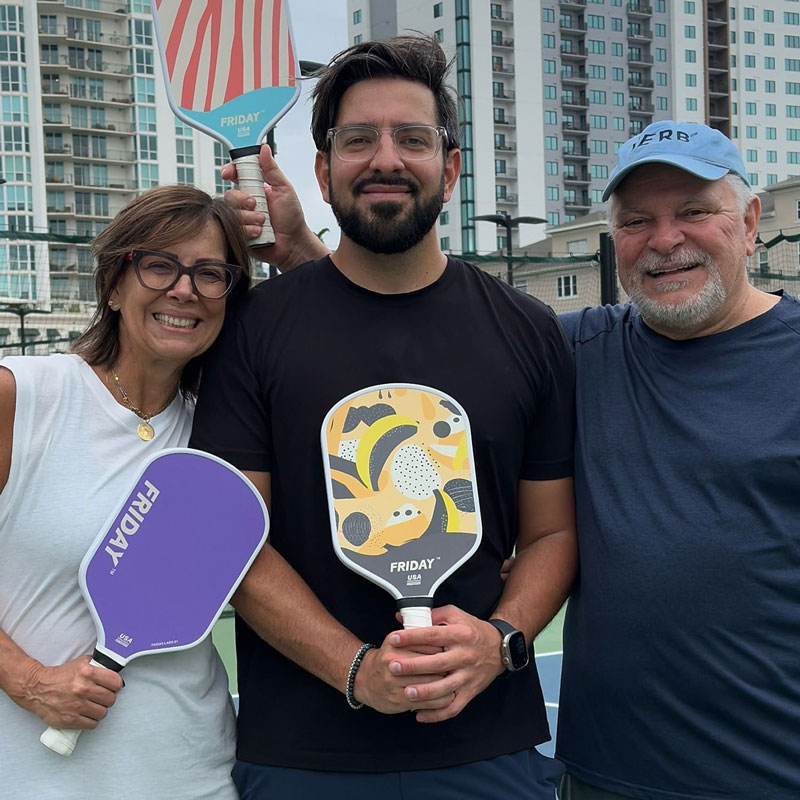Three people hold pickleball paddles on a court with buildings in the background.